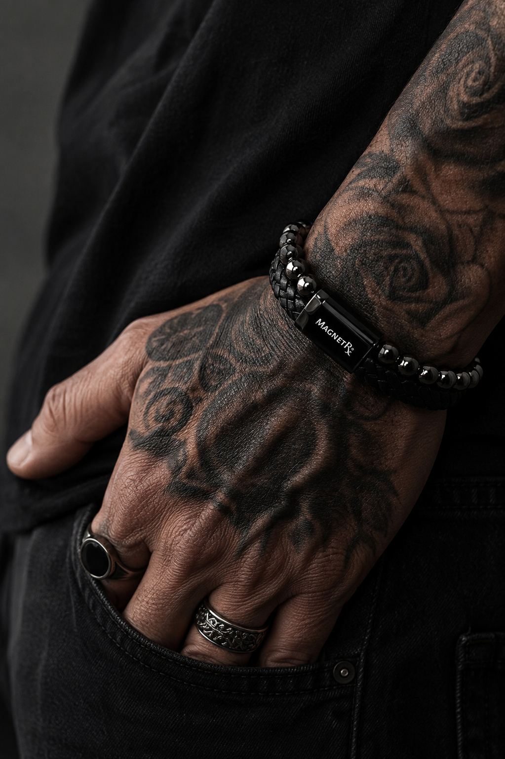 Tattooed man wearing a black braided leather bracelet with polished black beads and magnetic clasp in a dark, moody editorial setting.