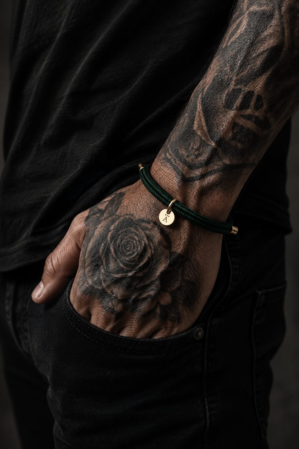 Tattooed man wearing a dark green braided rope bracelet with gold-tone accents and a small round charm, styled with an all-black outfit.