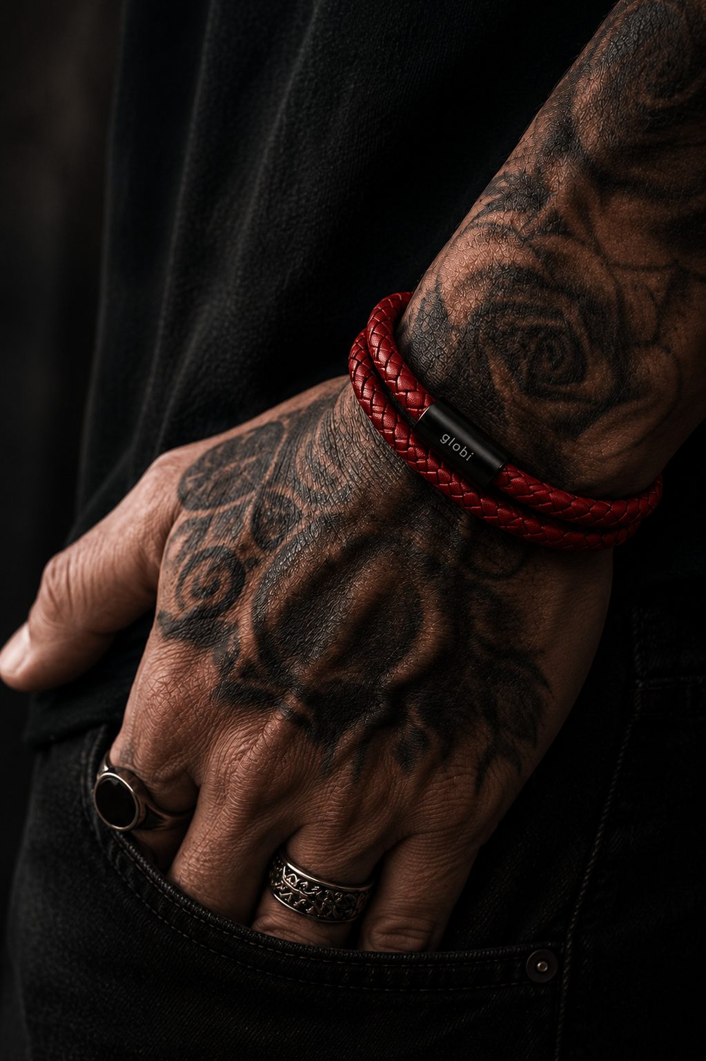 Close-up of a tattooed man wearing a red braided leather double-wrap bracelet with a black clasp, styled with black clothing and rings.