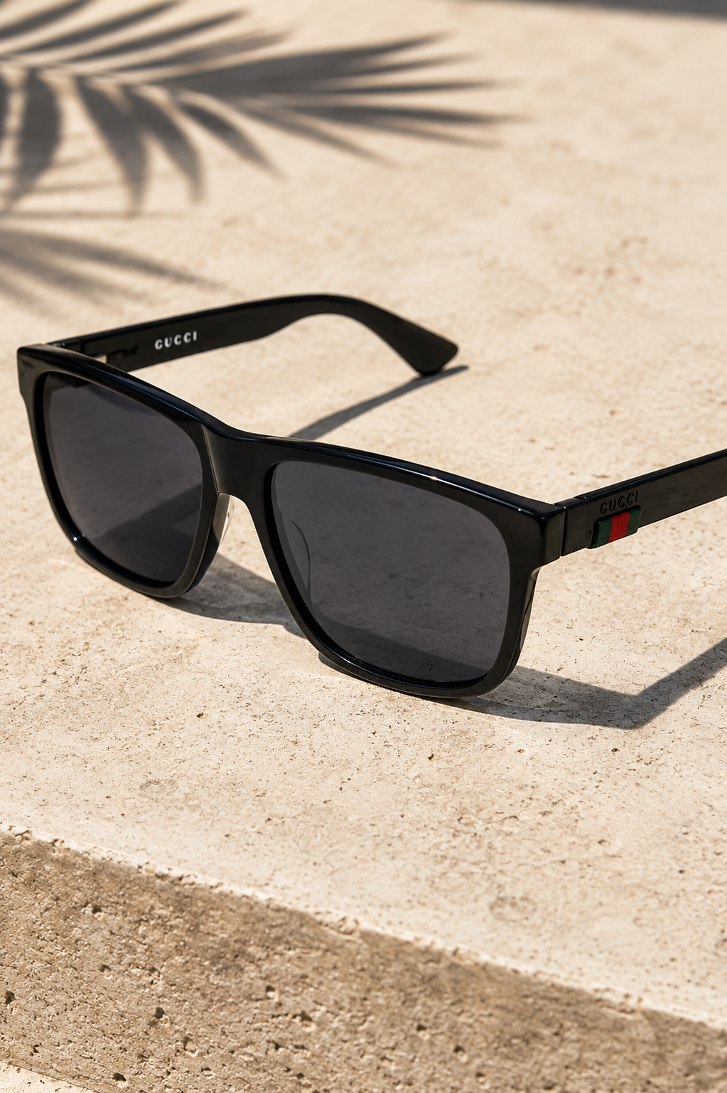 Black Gucci men's sunglasses with dark lenses and signature green and red temple detail displayed on a sunlit stone surface.