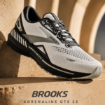 A Brooks Adrenaline GTS 23 men's running shoe in white, grey, and black resting on a textured, sandy-brown architectural ledge against an archway backdrop. Text at the bottom reads: "Brooks Adrenaline GTS 23. Cushion. Support. Confidence. Built for the long run."