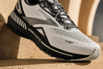 A Brooks Adrenaline GTS 23 men's running shoe in white, grey, and black resting on a textured, sandy-brown architectural ledge against an archway backdrop. Text at the bottom reads: "Brooks Adrenaline GTS 23. Cushion. Support. Confidence. Built for the long run."