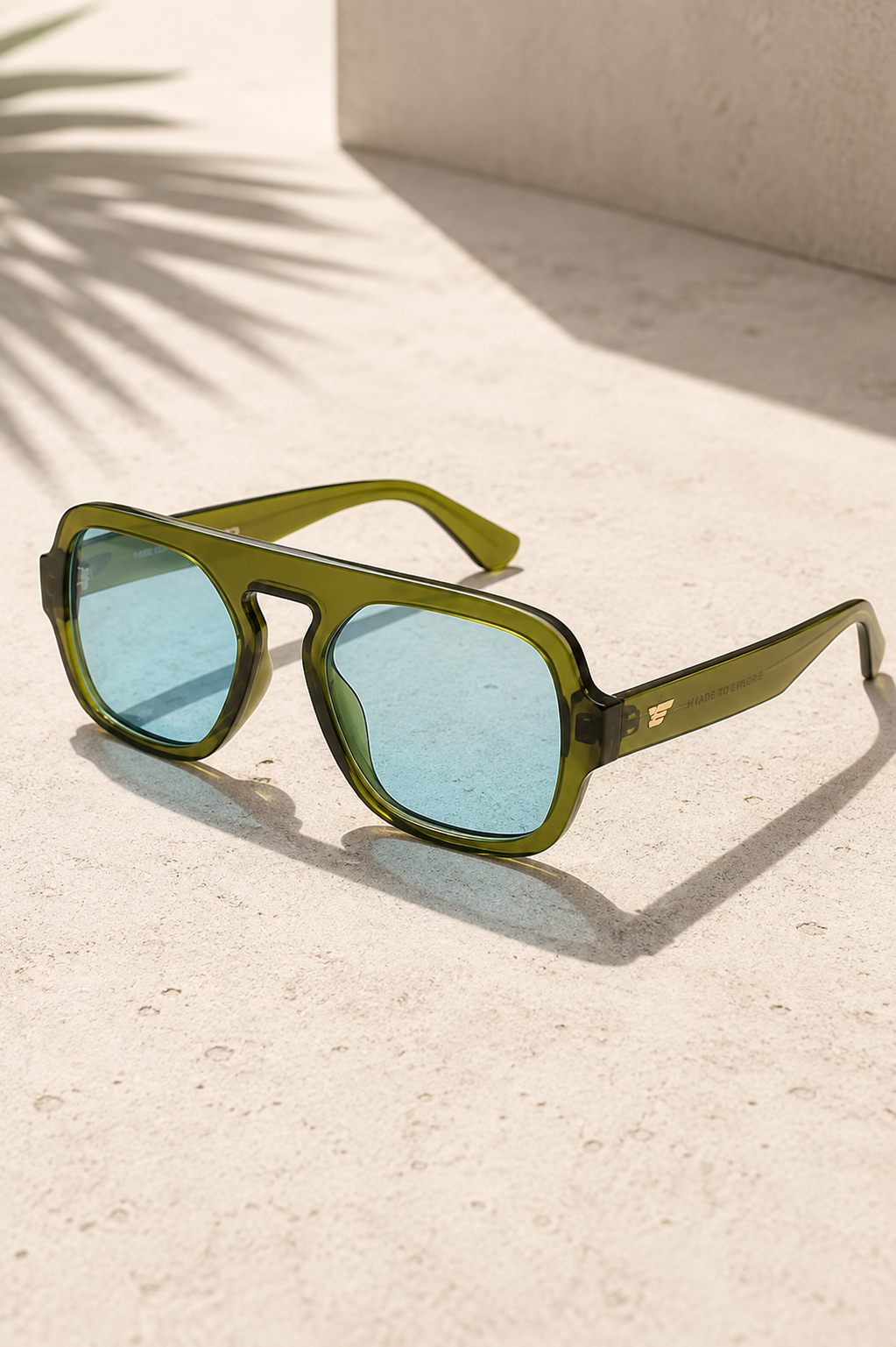 Green translucent men's sunglasses with light blue lenses displayed on a sunlit stone surface.