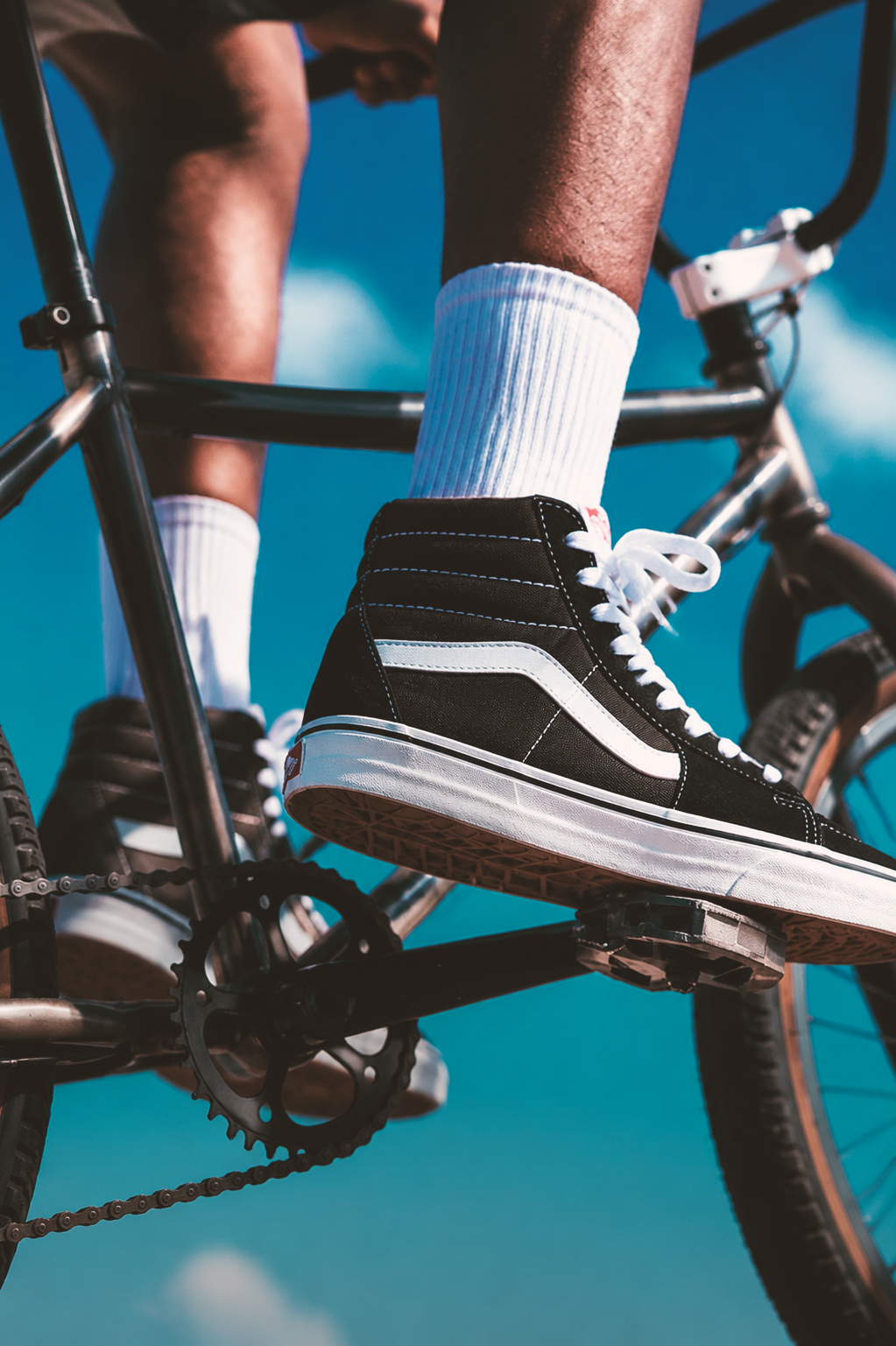 A close-up action shot of a person riding a BMX bike, wearing black and white high-top sneakers with white socks, focused on the feet and bike pedals against a bright blue sky.
