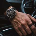 A close-up shot of a man's hand resting on a car gear shifter, wearing a luxury skeleton chronograph watch and a simple dark ring inside a modern vehicle.