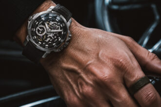 A close-up shot of a man's hand resting on a car gear shifter, wearing a luxury skeleton chronograph watch and a simple dark ring inside a modern vehicle.