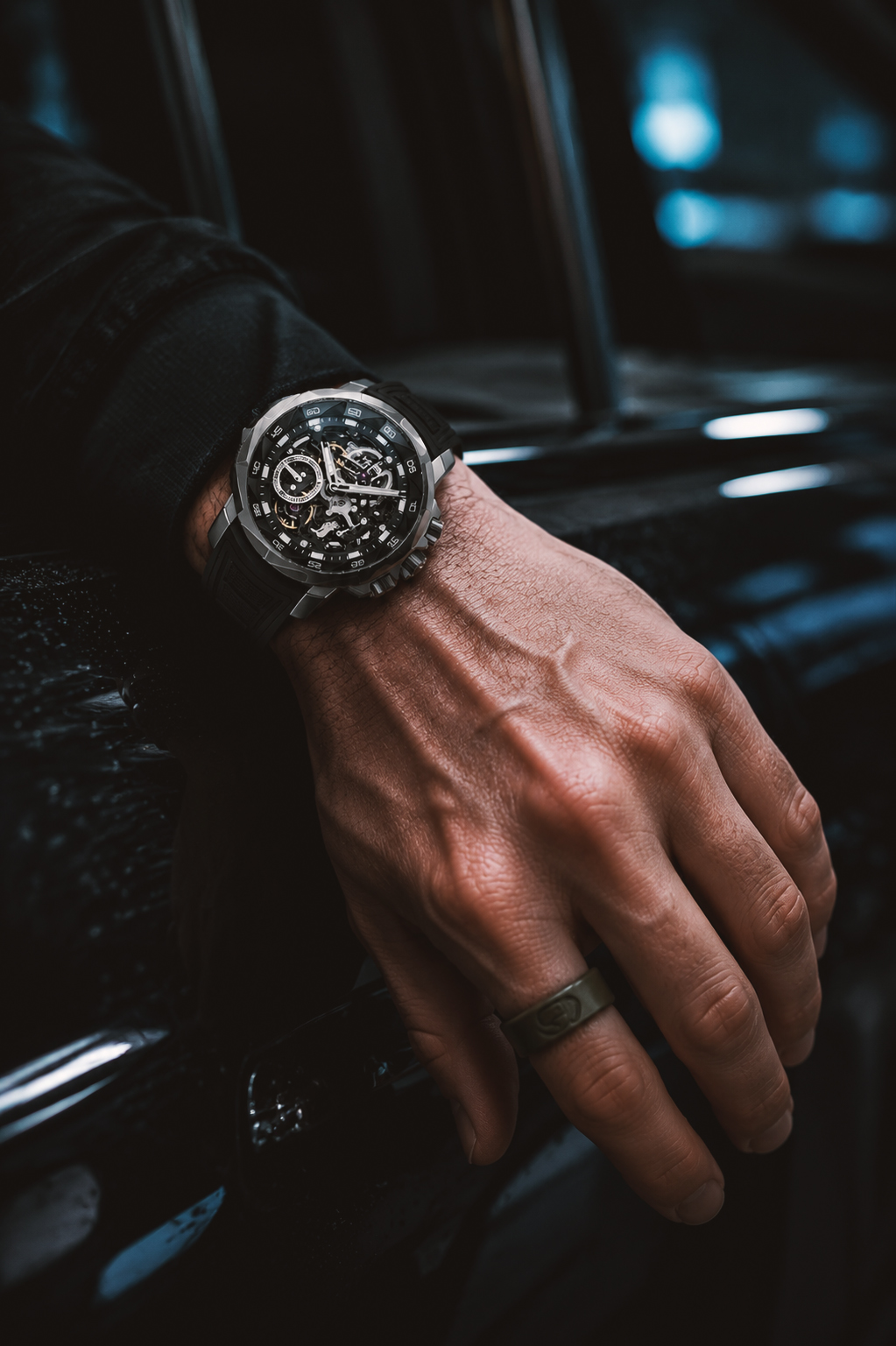 A close-up shot of a man's hand resting on a car gear shifter, wearing a luxury skeleton chronograph watch and a simple dark ring inside a modern vehicle.