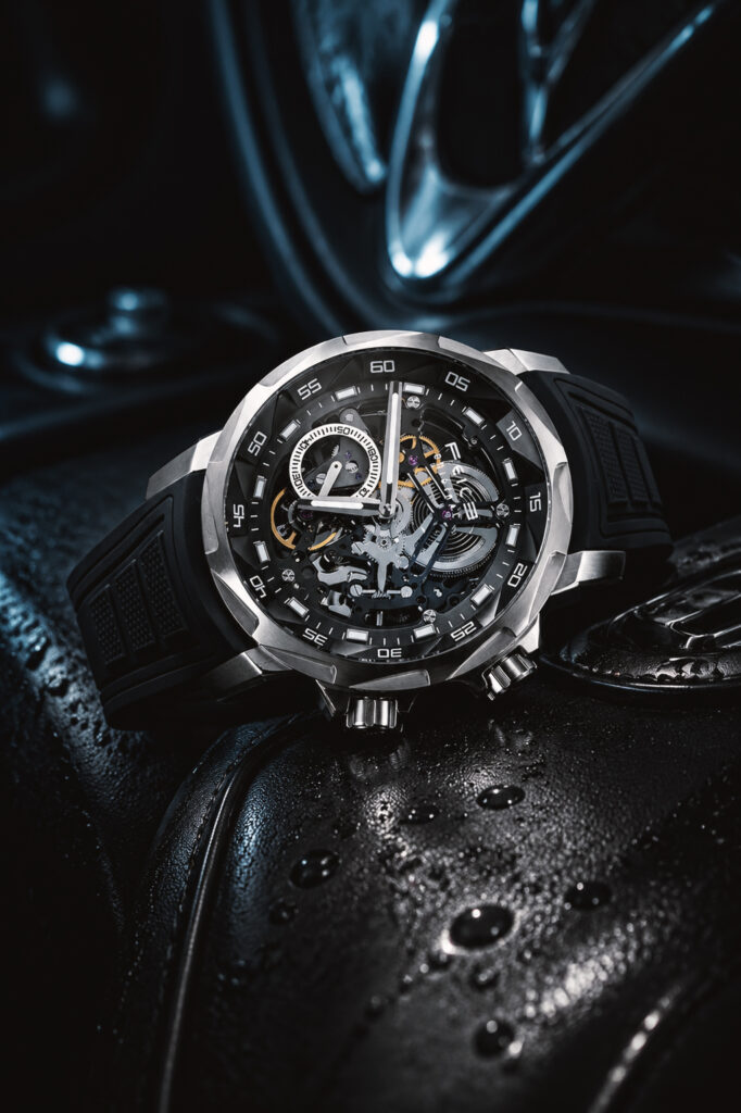 A close-up product shot of a luxury men's skeleton dial watch with a silver case and black band, resting on a dark, wet leather surface.