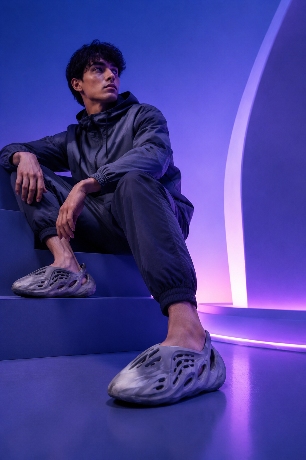 A young man sitting on a blue studio block against a vibrant purple and blue-lit background, wearing a dark hooded windbreaker, dark joggers, and marbled grey molded foam slip-on shoes.