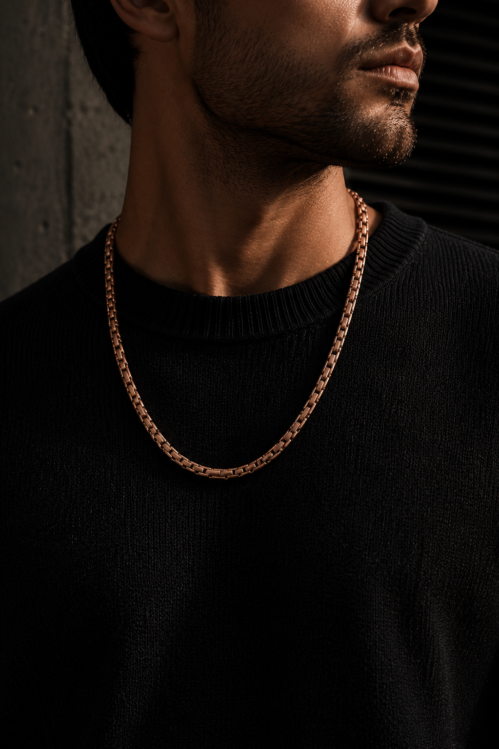 Man wearing a rose-gold magnetic necklace with a black outfit in a moody architectural setting.